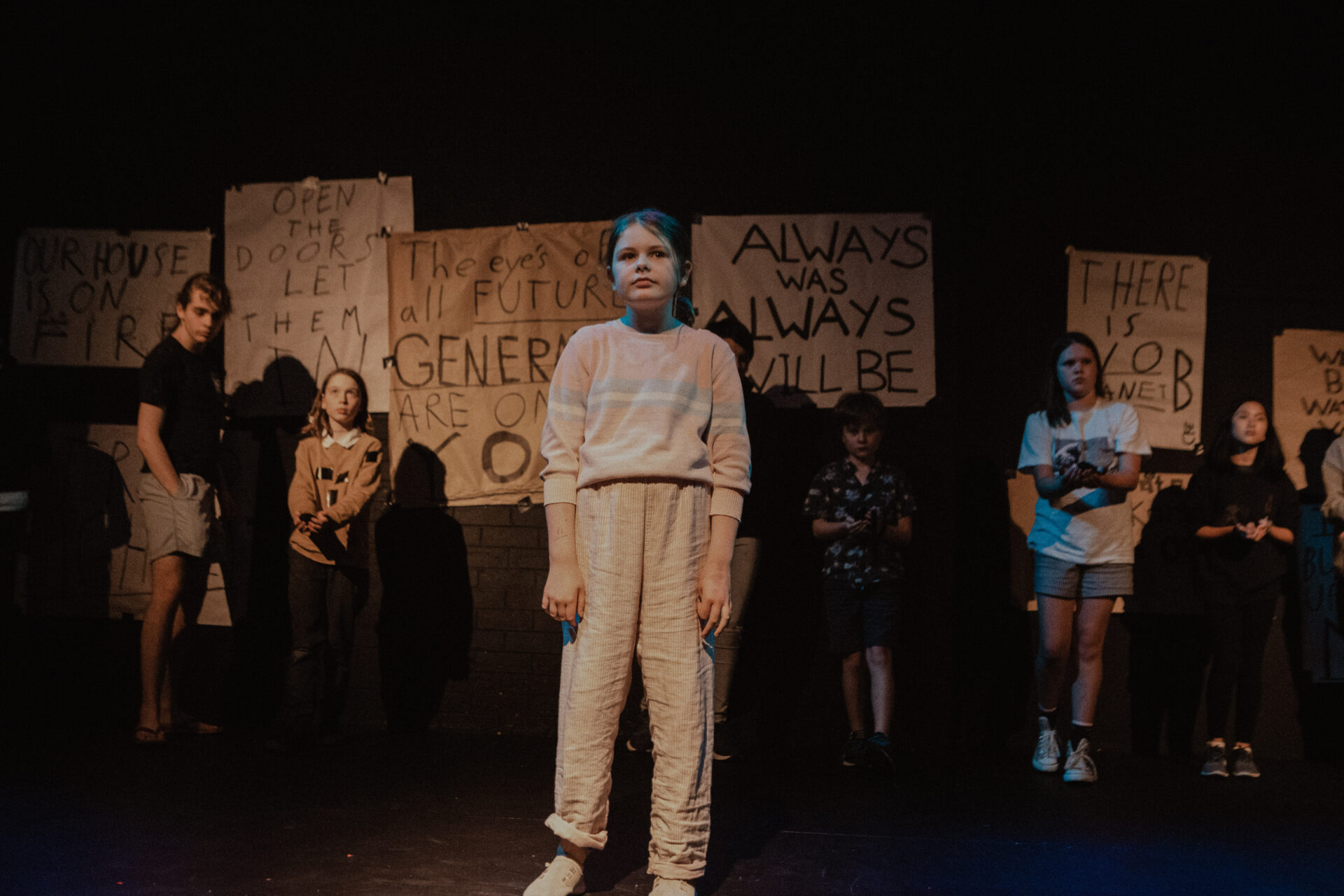 Young girl in white infront of crowd of young people and poster paper with words on them all staring at an audience under light