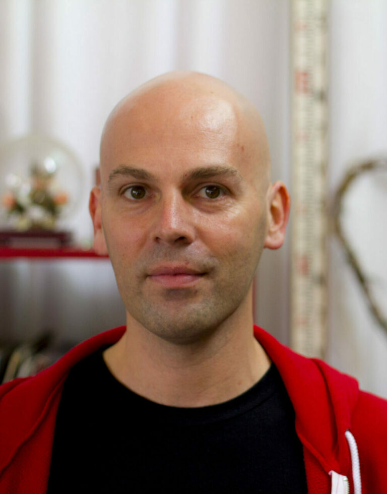 Daniel Tobias. A White man with a shaved head. He is looking to camera with a neutral expression. He is wearing a black t-shirt with a red zip up hoodie.