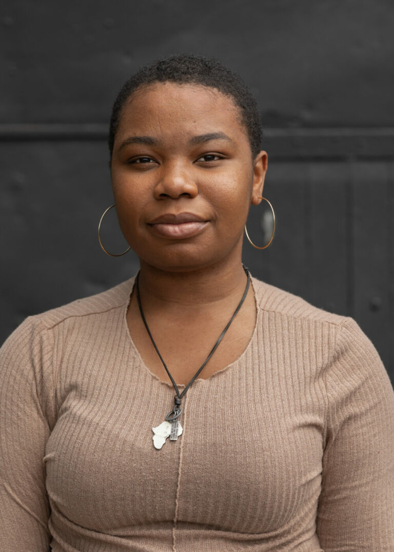 young igbo woman with shaved black hair, silver hoop earrings, tan shirt and necklace