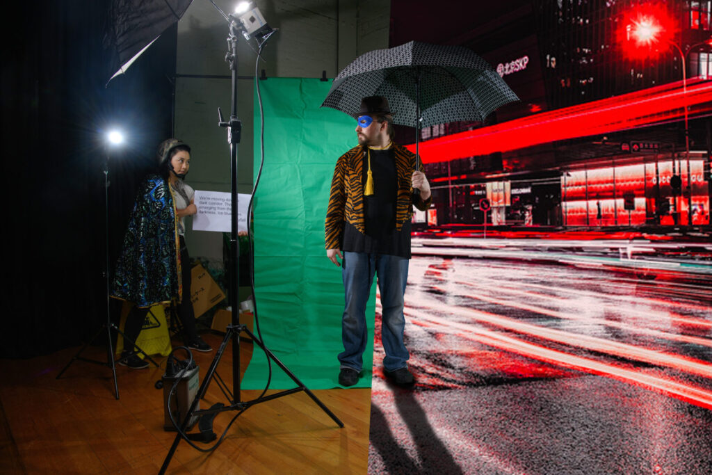 A person stands with an umbrella, centre, left side is a green screen and right side, a city street