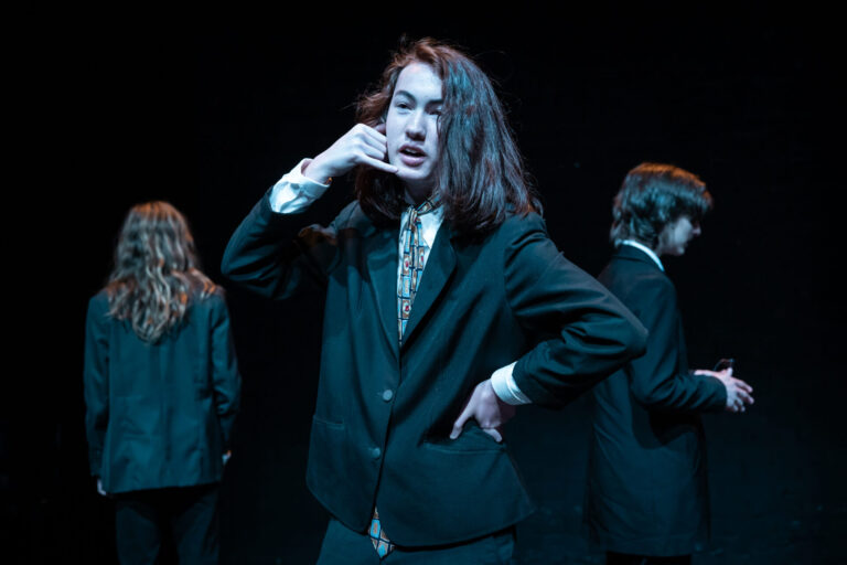 Three teenage performers stand on stage. They are wearing black suits. The one standing in the middle has one hand on his hip, the other mimicking a phone call. The other two are standing either side of him a little further back. The one on the left has their back to the camera, the one on the right is facing to the right of the frame and is holding their hands together as if in thought. The background is black.