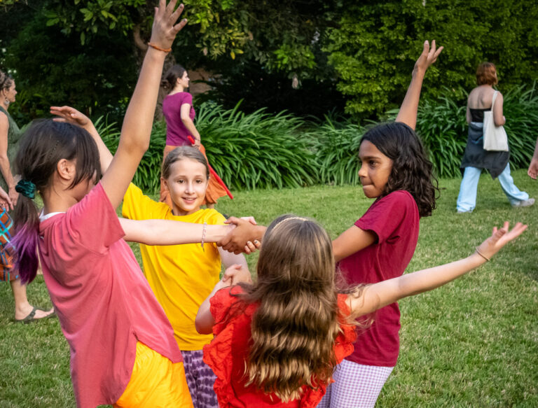 4 young girls in coloured t-shirts form a circle on the grass. They are holding each other's arms in the centre with their other arms leaning out of the circle (from Gene Tree Darwin performance)
