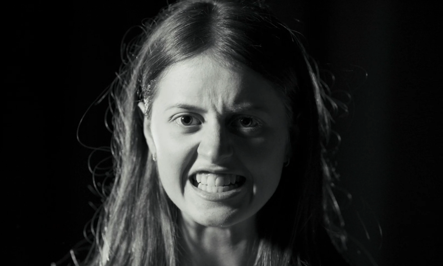 Black and white close up of teen girl gritting teeth, fiercely staring at camera.